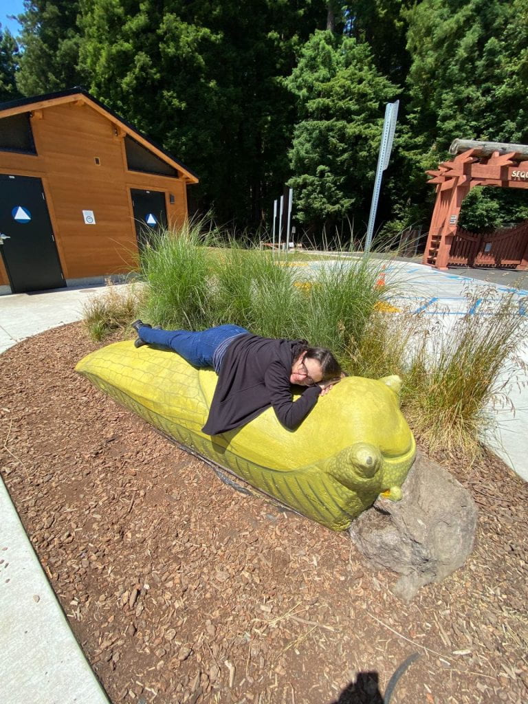 This is me, a white woman with dark hair wearing jeans and a black sweatshirt, laying on a larger-than-me cement, yellow banana slug. It's in a park.